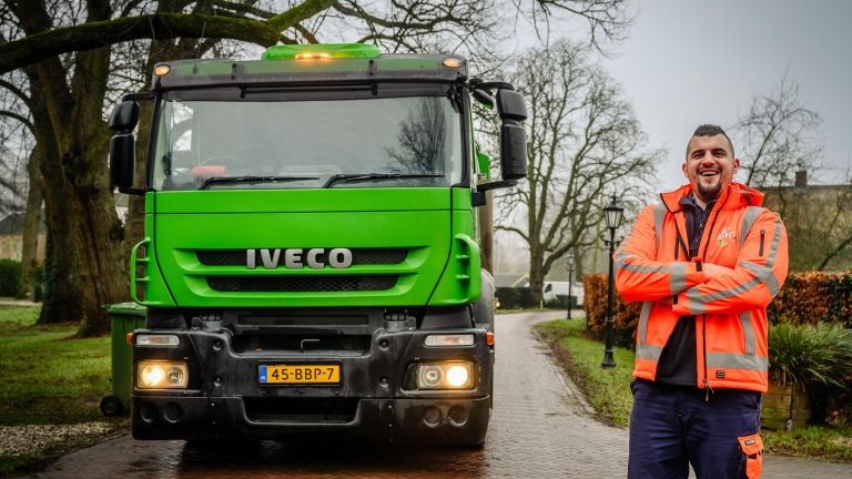 Avri chauffeur staat lachend met armen over elkaar voor een Avri inzamelwagen op een straat.