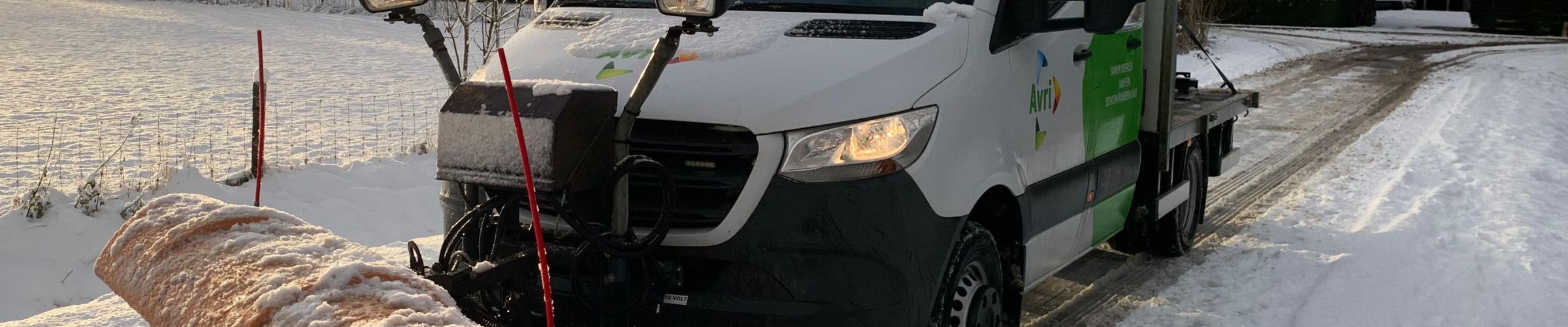 Avri-voertuig met sneeuwschuif maakt een besneeuwde landweg vrij in een winterse omgeving, met bomen en velden op de achtergrond.