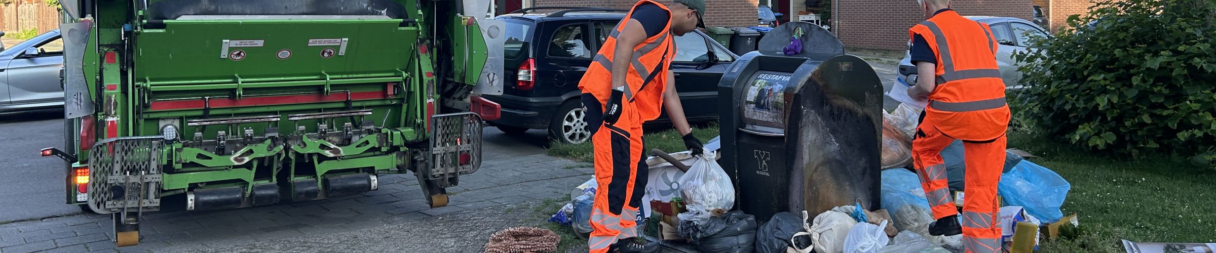 Een Avri vuilniswagen staat geparkeerd naast een ondergrondse afvalcontainer in een woonwijk. Twee Avri medewerkers in opvallende veiligheidskleding ruimen afvalzakken en los afval op dat naast de container is geplaatst.