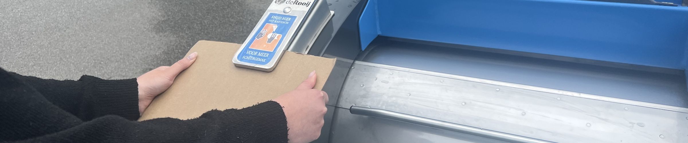 Persoon houdt een stuk karton vast en schuift deze onder de kartonsnijder van de ondergrondse papiercontainer.