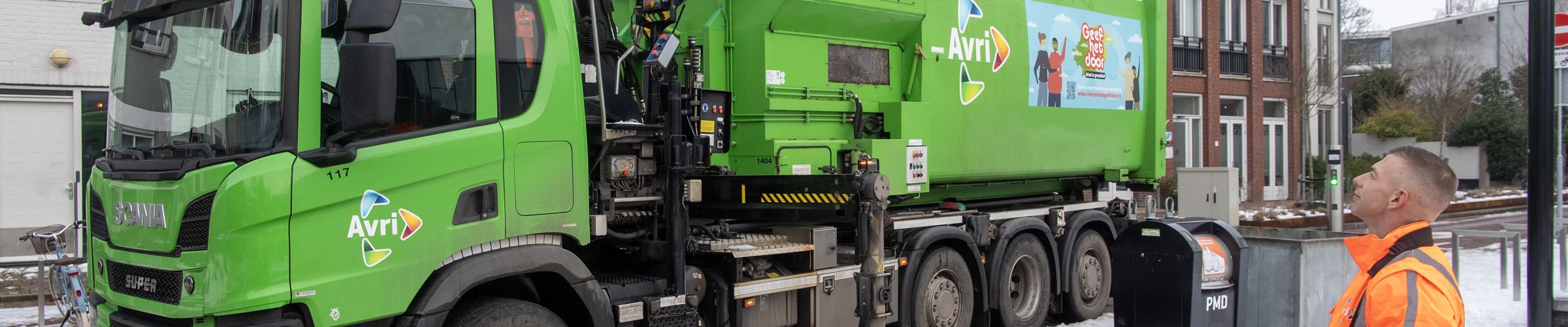 Een groene Avri-inzamelwagen leegt met behulp van een kraan een ondergrondse afvalcontainer. Op de voorgrond bedient een medewerker in opvallende veiligheidskleding de kraan met een afstandsbediening. Rondom de container ligt sneeuw.
