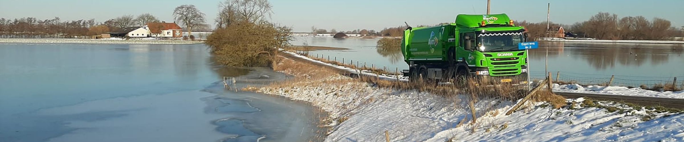 Groene inzamelwagen rijdt over een besneeuwd dijkje langs een bevroren rivier in een landelijk winterlandschap.