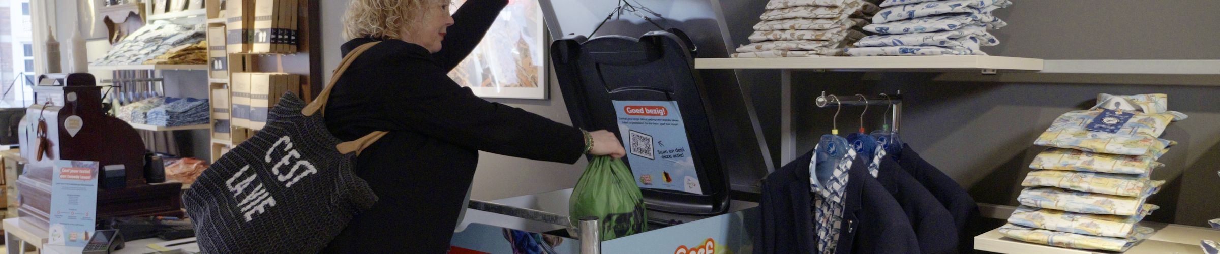 Een persoon staat in een winkel en deponeert een zak textiel in een textielinzamelcontainer. De container is kleurrijk bedrukt met illustraties en teksten. De ruimte bevat kledingrekken met opgevouwen kleding, een toonbank met diverse artikelen en decoratieve schilderijen aan de muur.