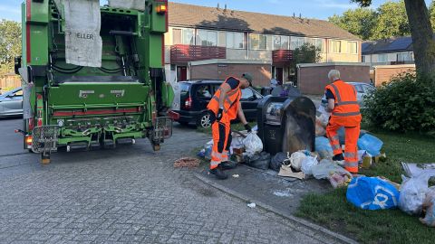 Een Avri vuilniswagen staat geparkeerd naast een ondergrondse afvalcontainer in een woonwijk. Twee Avri medewerkers in opvallende veiligheidskleding ruimen afvalzakken en los afval op dat naast de container is geplaatst.
