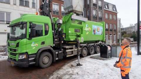 Een groene Avri-inzamelwagen leegt met behulp van een kraan een ondergrondse afvalcontainer. Op de voorgrond bedient een medewerker in opvallende veiligheidskleding de kraan met een afstandsbediening. Rondom de container ligt sneeuw.